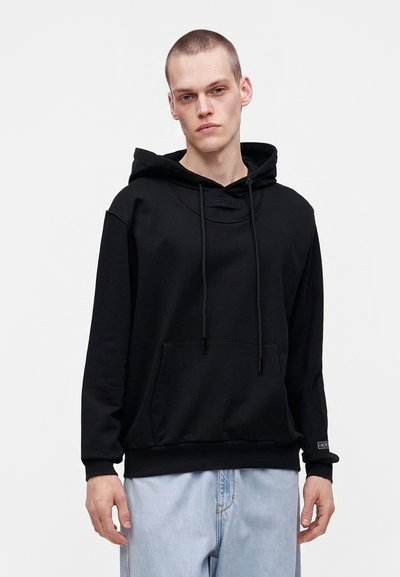 Jeune homme aux cheveux courts portant un sweat à capuche noir et un jean bleu clair, debout devant un fond blanc uni.