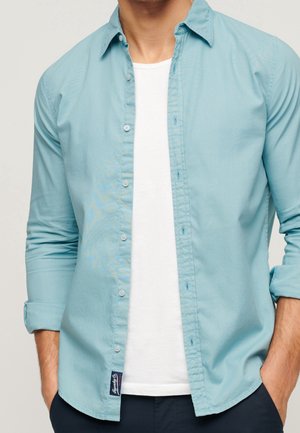 Mężczyzna ubrany w jasnoniebieską koszulę na guziki na białej koszulce z podwiniętymi rękawami, ręce w kieszeniach ciemnych spodni, widoczny tors.