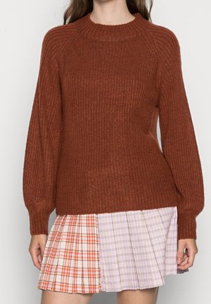 Femme portant un pull en maille côtelée couleur rouille et une mini-jupe plissée avec des motifs à carreaux orange et violet.