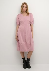 Rosa bomullsklänning med korta puffärmar, V-ringning och knälång silhuett. Bärs med svarta ankelstövlar. Slät textur och enkel design.