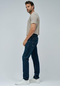 Hombre de pie de perfil vistiendo una camiseta beige clara, jeans azul oscuro y zapatillas blancas frente a un fondo gris claro liso.