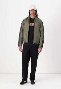 Grön bomberjacka med frontdragkedja, svarta ränder på ärmarna, bärs över en svart t-shirt med grafik och matchas med svarta byxor och sneakers.