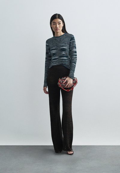 Kvinna i långärmad blåmönstrad topp, genomskinliga svarta byxor, svarta öppna klackskor, hållande en röd och vit mönstrad kuvertväska med kedja.