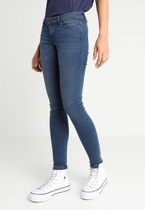 Personne portant un jean skinny bleu et des baskets montantes blanches, debout devant un fond clair uni.