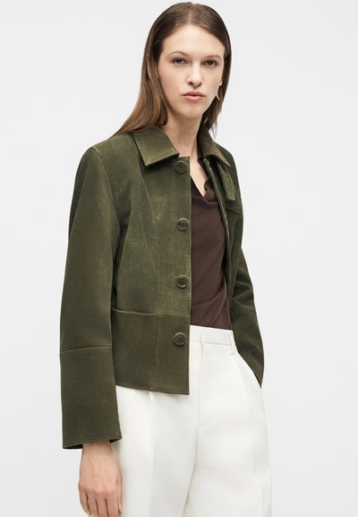 Γυναίκα που φοράει μια σακάκι από πράσινο σαμουά με κουμπιά, ένα καφέ τοπ και ψηλόμεσα λευκά παντελόνια, στέκεται μπροστά σε απλό φόντο.