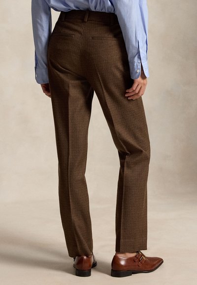 Homme portant un pantalon sur mesure à carreaux marron, une chemise habillée bleu clair et des chaussures en cuir marron à double boucle, debout devant un fond uni.