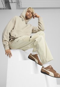 Felpa beige con mezzo zip e cappuccio attaccato, pantaloni cargo crema e sneakers Puma marroni con dettagli bianchi. Ambientato su uno sfondo grigio chiaro.