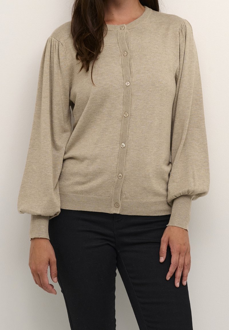 Femme portant un cardigan beige boutonné avec des manches longues bouffantes et un pantalon noir, debout devant un fond clair uni.