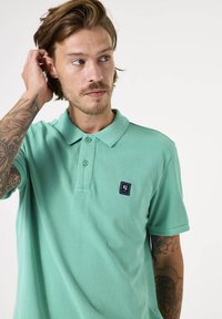 Polo de color verde menta fabricado en algodón, con una tapeta de dos botones y un pequeño parche de logo en el lado izquierdo del pecho. Mangas cortas.