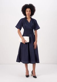 Vestido azul marinho até ao joelho, com mangas curtinhas, apresenta uma cintura franzida e detalhes em costura branca. Modelo segurando uma carteira branca.