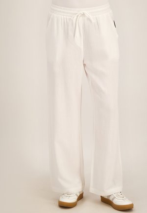 Persoon met losse witte broek met trekkoord en witte sneakers met lichtbruine zolen, staand tegen een egale lichte achtergrond.