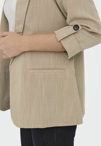 Beige blazer van textuurstof, met een getailleerde pasvorm, een enkele borstzak en een contrasterende zwarte knoop op de opgerolde mouw.