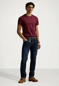 T-shirt de gola redonda bordô, jeans de ganga azul, cinto castanho com listra verde e mocassins castanho-claro usados por um modelo masculino em pé.