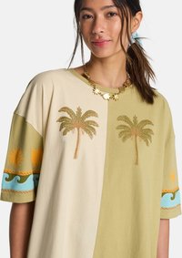 Chemise oversize bicolore beige et vert olive avec broderie de palmier. Elle présente un col rond et des détails de manches vibrants.