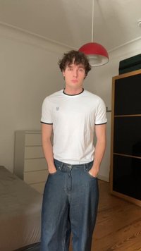 Hvit T-skjorte med kort erm og svart kant, med en liten logo, sammen med løse blå denimjeans. Bakgrunnen har lyst tregulv.