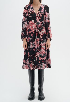 Femme portant une robe midi noire avec des motifs floraux rouges et roses et des manches longues, associée à des bottes en cuir noir jusqu'au genou.