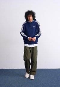 Donkerblauwe hoodie met witte strepen en logo, gelaagd over een wit shirt, gecombineerd met olijfgroene cargo-broeken en witte sneakers op een blauwe vloer.