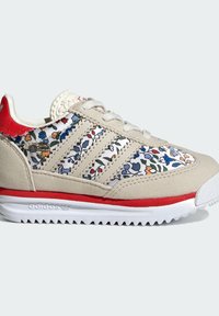 Beige children's sneaker with floral pattern, red accents, and three white stripes. Made of suede with a textured rubber sole.
