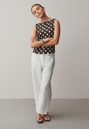 Femeie stând cu brațele încrucișate, purtând un top negru fără mâneci cu buline albe, pantaloni albi, sandale negre și accesorii argintii.