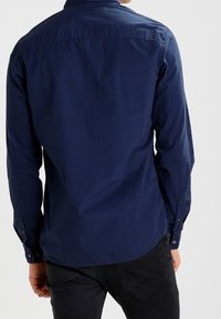 Laivastonsininen pitkähihaisa paita, joka on valmistettu kudotusta kangasta; siinä on kaulus, nappi- ja hihansuut sekä kaareva helma. Ei näkyviä kuvioita tai logoja.