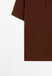 Polo marron à manches courtes en tissu texturé avec une patte boutonnée visible sur fond blanc.