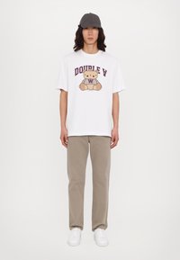 Camiseta blanca con un gráfico de un oso de dibujos animados y el texto "DOUBLE Y" en púrpura. Combinada con pantalones beige claros y zapatillas blancas, rematada con una gorra gris.