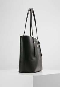 Czarna torebka tote z sztucznej skóry o gładkiej fakturze, z dwoma uchwytami i metalicznym okrągłym akcentem z przodu. Prostokątny kształt i pojemna.