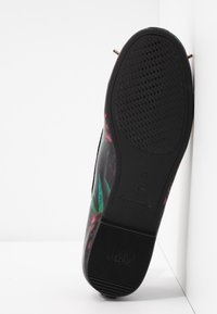 Semelle de chaussure plate noire avec grip texturé, motif floral latéral, taille 37, et étiquette antidérapante positionnée verticalement contre un mur blanc.