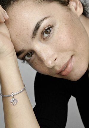 Silberne Armbanduhr mit einem strukturierten Band und einem herzförmigen Anhänger, der kunstvolle Ausschnitt-Designs aufweist. Das Handgelenk des Modells ist teilweise sichtbar.