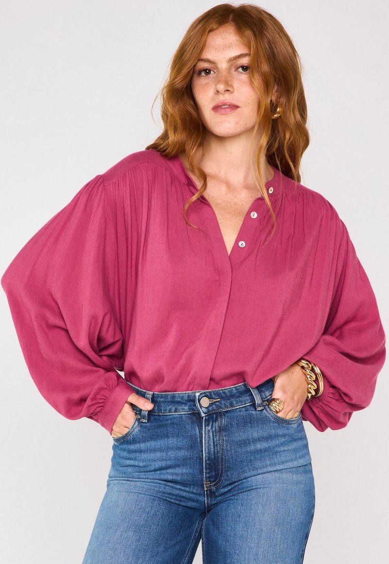 Blusa di un rosa intenso, con scollatura a V con bottoni, spalle plissettate e maniche lunghe ampie, abbinata a jeans in denim blu a vita alta.