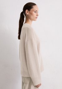 Beige oversized trui gemaakt van zacht materiaal, met een geribbelde zoom en manchetten, en een ontspannen pasvorm. Gecombineerd met lichtgekleurde broeken.
