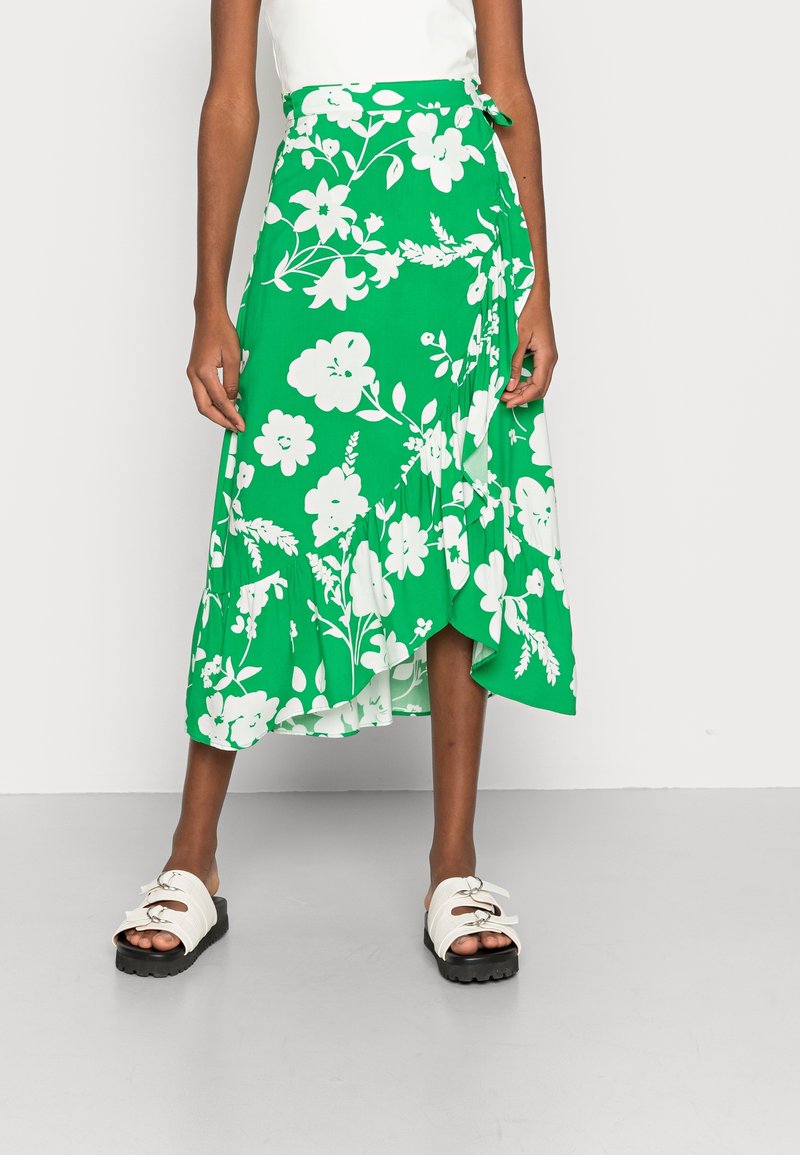 Jupe portefeuille verte à fleurs blanches, avec un ourlet évasé et un détail noué à la taille. Associée à des sandales blanches avec des semelles noires.