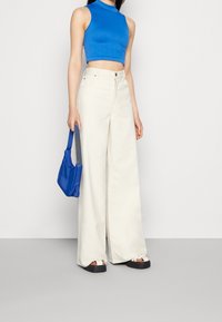 Top corto azzurro, pantaloni bianchi a gamba larga, sandali neri con piattaforma e una piccola borsa blu. L'outfit presenta linee semplici e pulite.