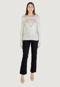 Suéter gris claro con un diseño de logo triangular, adornado con strass, combinado con pantalones negros ajustados y zapatos color nude.