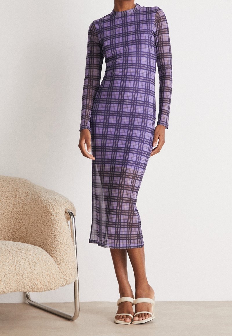 Robe à manches longues à carreaux violets, dotée d'une silhouette ajustée avec un tissu transparent. Le mannequin se tient à côté d'une chaise couleur crème aux pieds métalliques.