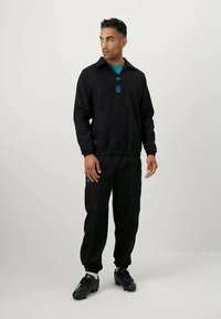 Juodas sportinis kostiumas su apykakle užsegamu švarku ir elastinga juostele ant kelnių. Krūtinėje ir kišenėse yra mėlynas logotipas, pagamintas iš lygaus audinio.