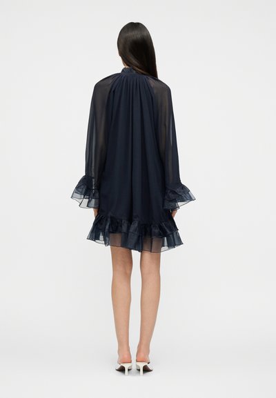 Γυναίκα με μακριά σκούρα μαλλιά που φοράει μπλε σκούρο διάφανο φόρεμα με βολάν στα μανίκια και το τελείωμα, στέκεται με την πλάτη, εμφανείς λευκές ψηλοτάκουνες γόβες.