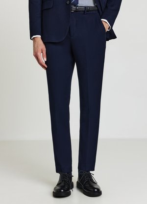 Homme portant un pantalon de costume bleu marine, un blazer assorti, une chemise rayée et des chaussures en cuir noires à lacets, debout avec une main dans la poche.