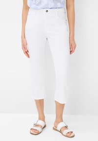 Pantalons blancs coupés en tissu lisse, avec une fermeture à un bouton et des poches avant, associés à des sandales blanches à enfiler.