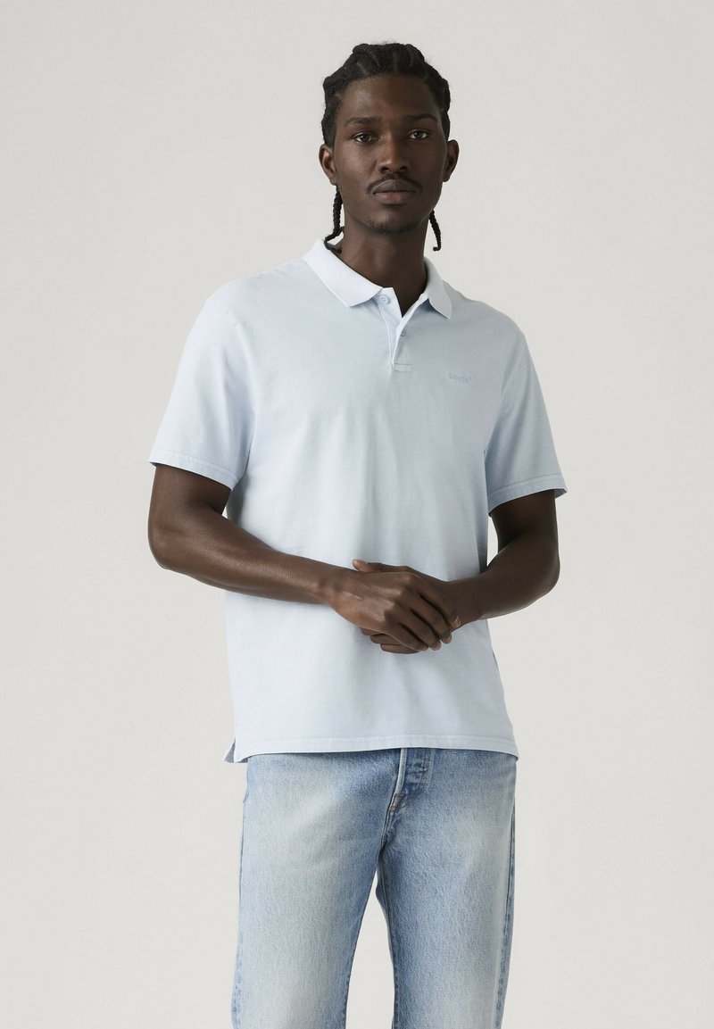 Hombre con cabello trenzado que lleva una camiseta tipo polo azul claro y jeans azules desgastados, de pie con las manos juntas contra un fondo liso.