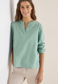 Jeune femme souriante, portant un chemisier à manches longues texturé vert menthe avec les poignets retroussés et un pantalon beige clair, appuyée contre un mur blanc.