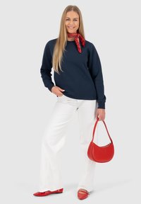 Marineblauwe sweatshirt, witte wijde jeans, rode platte schoenen en een bijpassende rode handtas. Sjaal met stippen geknoopt om de hals. Gladde texturen.