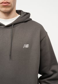 Άτομο που φοράει μια σκούρα γκρι μπλούζα φούτερ New Balance με κορδόνια, πάνω από λευκό μπλουζάκι με στρογγυλή λαιμόκοψη, ορατό από τους ώμους μέχρι το μέσο του στήθους.