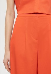 Orange wide-leg trousers with a smooth texture, featuring a single front pocket and a button closure at the waist. Simple and modern design.