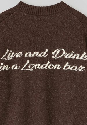 Pull marron avec un tricot texturé et un col côtelé. Présente un texte brodé en blanc : "Vis et Bois dans un bar londonien."