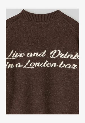 Pull marron avec un tricot texturé et un col côtelé. Présente un texte brodé en blanc : "Vis et Bois dans un bar londonien."