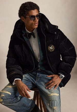 Jeune homme portant des lunettes de soleil, une doudoune noire avec un emblème, une chemise blanche avec un nœud papillon et un jean bleu déchiré, assis sur un tabouret en bois.