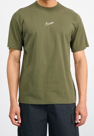 Homme portant un t-shirt Nike à manches courtes vert olive avec un petit logo blanc Swoosh sur la poitrine et un short noir avec poche latérale.