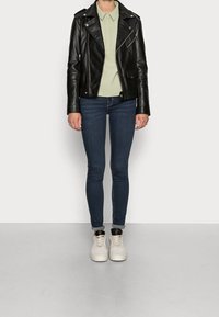 Persona vestida con una chaqueta de cuero negra, una camisa de cuello verde claro, jeans ajustados oscuros y zapatillas blancas, de pie contra un fondo gris.