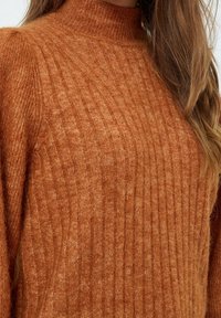 Primer plano de una persona que lleva un suéter de punto acanalado en color óxido, con un cuello alto y mangas largas.